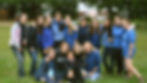 A group of 15 smiling people in blue uniforms pose on a grassy lawn with trees and a dock in the background.