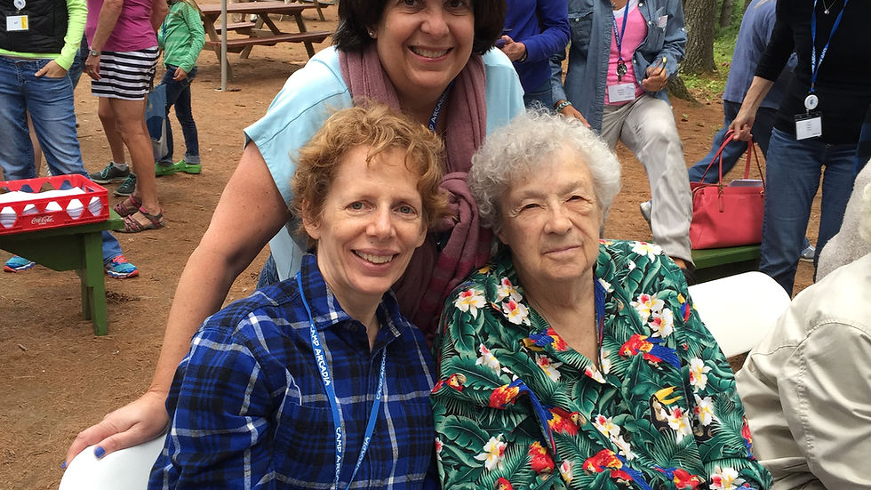 Three women smiling in a forest setting with a tent. One wears a floral shirt and holds a cane. Others wear blue shirts. Mood is cheerful.