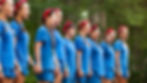 Girls in blue shirts and red bandanas sing outdoors, linked hand-in-hand. Trees in the background create a lively and cheerful scene.