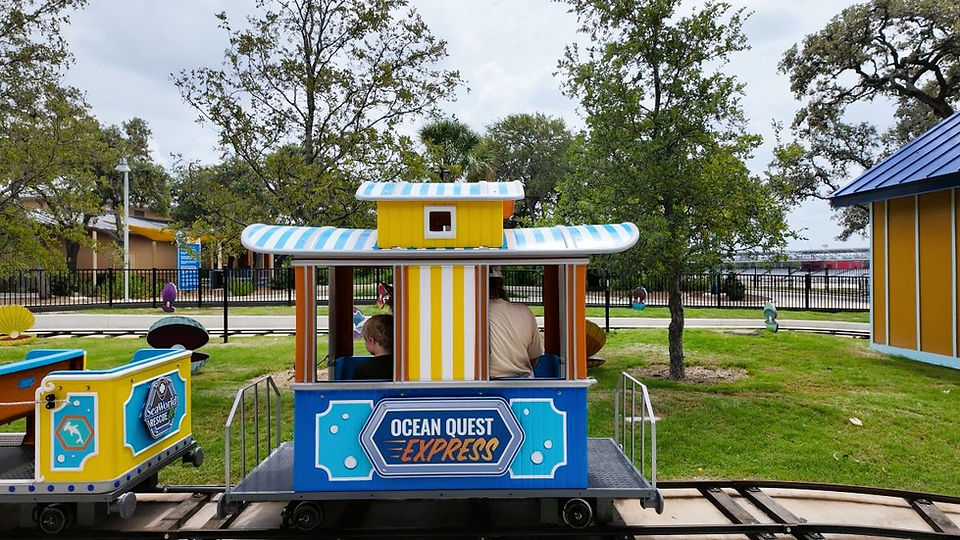 Kids ride a colorful "Ocean Quest Express" train in a park setting with green grass and trees. The train features bright blue and yellow hues.