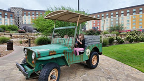Two children in a green vintage Jeep playfully pretending to drive. They're in a garden setting with elephant statues and a colorful hotel.