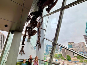 Children standing beneath a dinosaur skeleton overlooking the Dallas skyline at the Perot Museum.