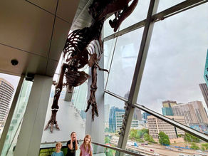 Children standing beneath a dinosaur skeleton overlooking the Dallas skyline at the Perot Museum.