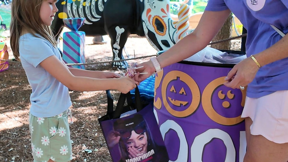 Girl receiving candy in a purple bag with Halloween motifs. Background shows a colorful skeleton display, trees, and wood chips.