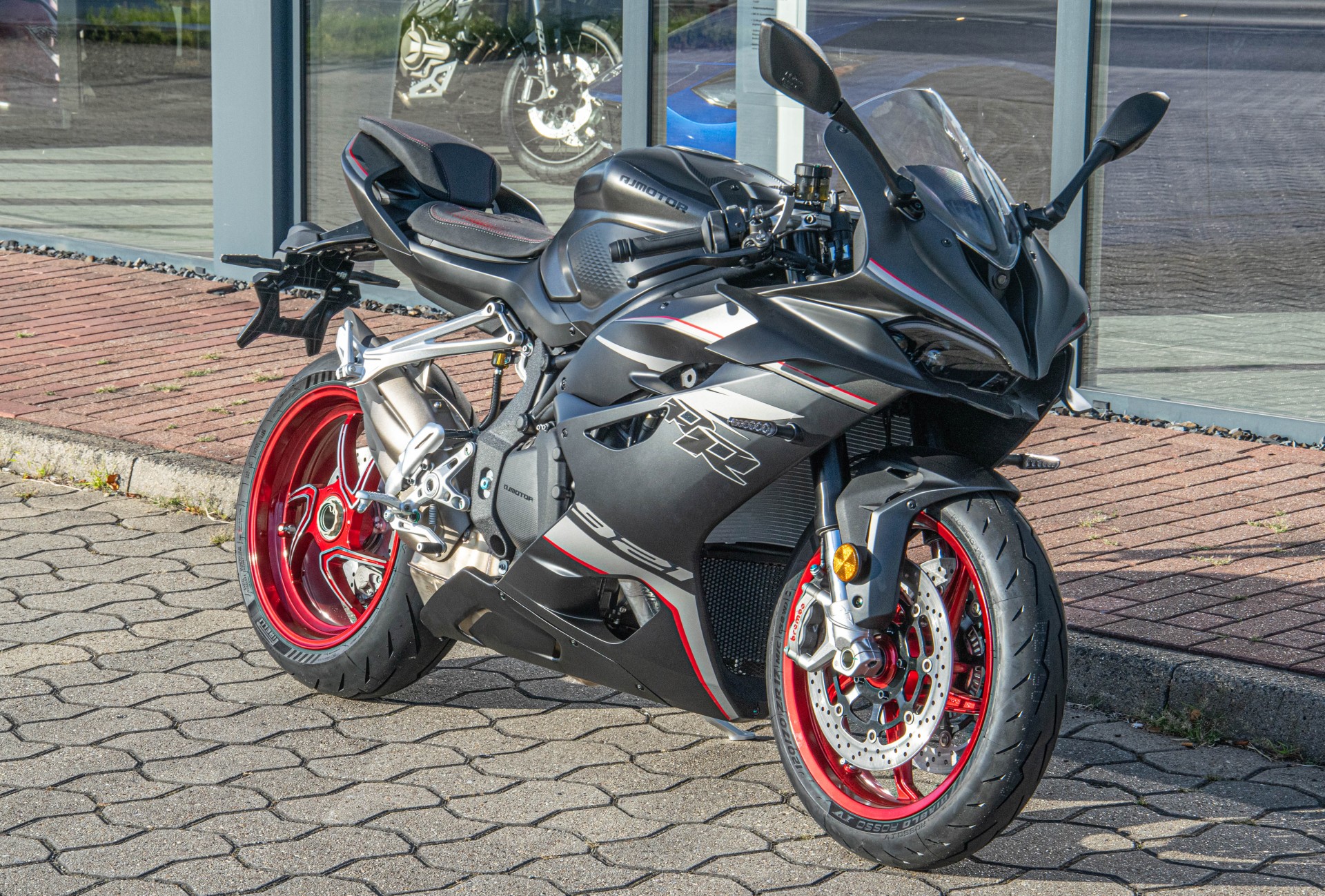 Schwarz-grauer QJMOTOR SRK 921 RR mit flammenartigen Grafiken und roten Felgen, vollständig ohne Fahrer auf Pflaster geparkt. Die seitliche Dreiviertelansicht zeigt die Vollverkleidung; hinter dem Motorrad liegt die Glasfront eines Showrooms.