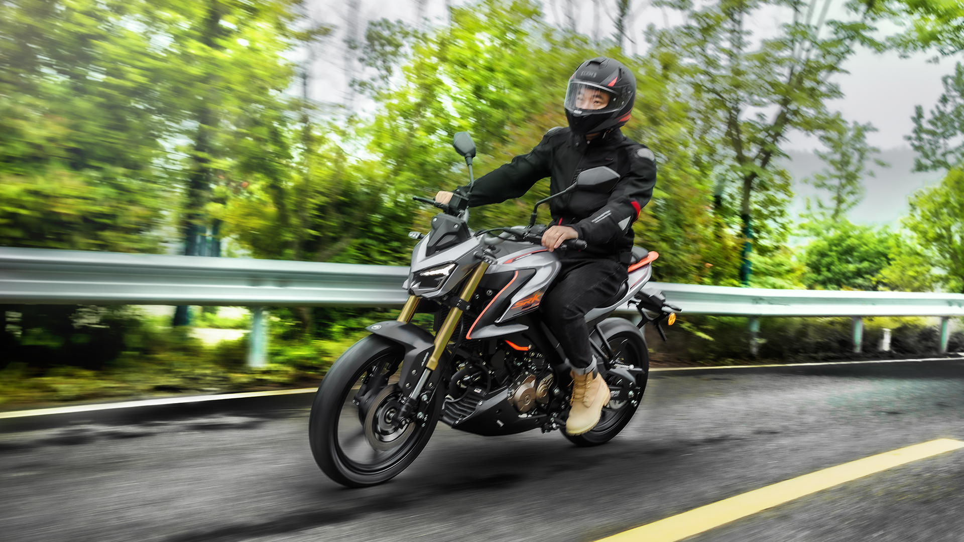 Dunkler QJMOTOR SRK 125 S in Schräglage auf einer kurvigen Landstraße. Die Person trägt einen geschlossenen Helm und dunkle Motorradkleidung; grüne Bäume, Leitplanke und Bewegungsunschärfe im Hintergrund vermitteln die Fahrt.