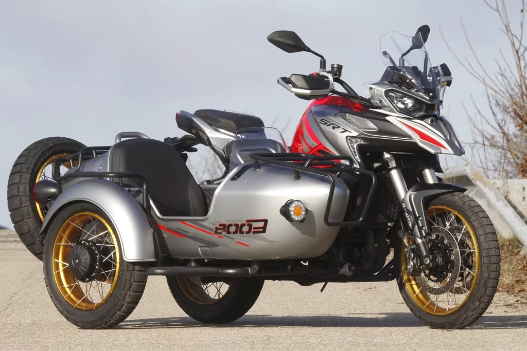 QJMOTOR SFT 900 mit rotem Tank, silberfarbenem Beiwagen und goldfarbenen Rädern, vollständig ohne Fahrer auf einer Landstraße geparkt. Alle drei Räder sind sichtbar; kahle Sträucher, ein braunes Feld und ein blauer Himmel mit Wolken bilden den Hintergrund.
