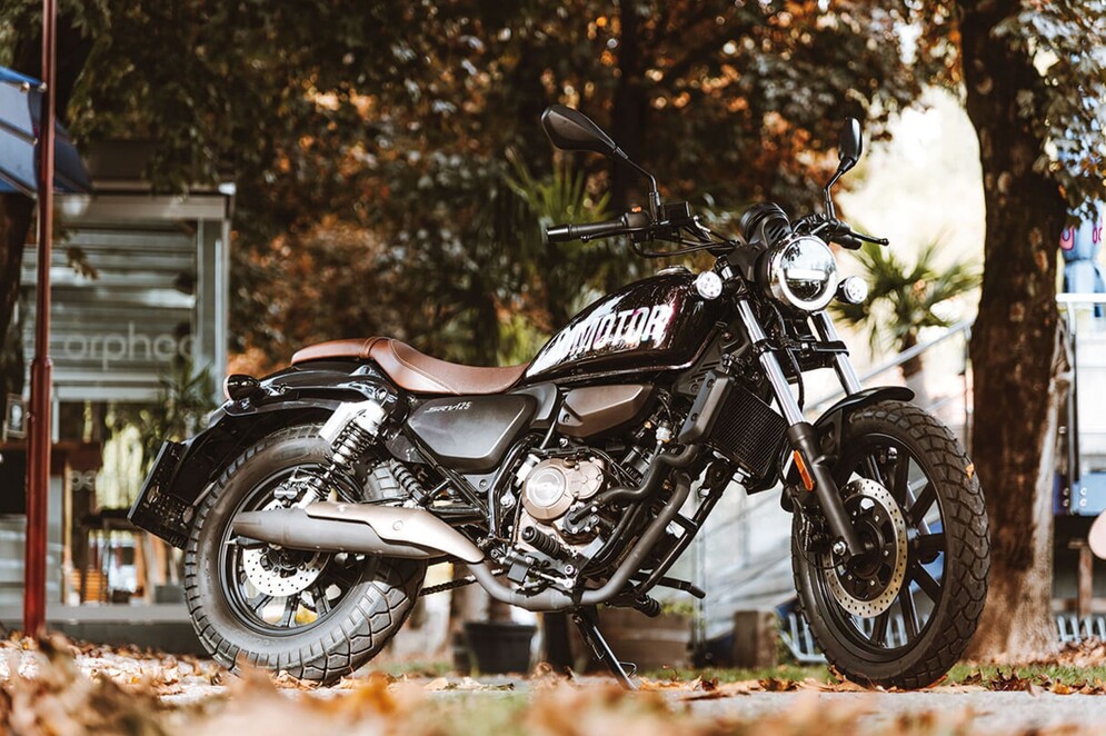 Schwarzer QJMOTOR SRV 125 mit brauner Sitzbank, vollständig vor einem Dorf-Café unter einem herbstlich gefärbten Baum geparkt. Motor und glänzender Auspuff sind in der Seitenansicht sichtbar; auf oder vor dem Motorrad befindet sich keine Person.