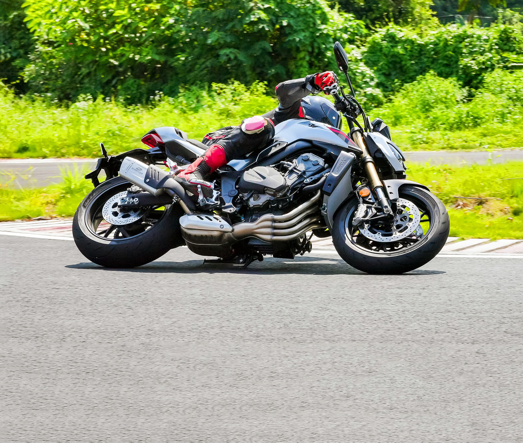 Grau-schwarzer QJMOTOR SRK 800 während einer Kurvenfahrt auf Asphalt, vollständig von der Seite aufgenommen. Die Person trägt schwarzen Helm und rot-schwarze Motorradkleidung; grüne Straßenränder und bewaldete Hänge bilden den Hintergrund.
