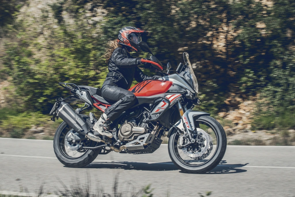 Rot-grauer QJMOTOR SRT 600 SX während der Fahrt auf einer kurvigen, von Wald umgebenen Asphaltstraße. Die Person trägt schwarzen Helm und dunkle Motorradkleidung; das Adventure-Motorrad mit hoher Scheibe und Speichenrädern ist vollständig von der Seite sichtbar.