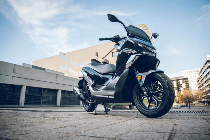 Grauer QJMOTOR FORT 125 N, ohne Fahrer in einer niedrigen Dreiviertelansicht auf einem gepflasterten Platz geparkt. Der Roller ist einschließlich beider Räder sichtbar; dahinter stehen moderne Gebäude mit Ziegel- und Betonflächen.