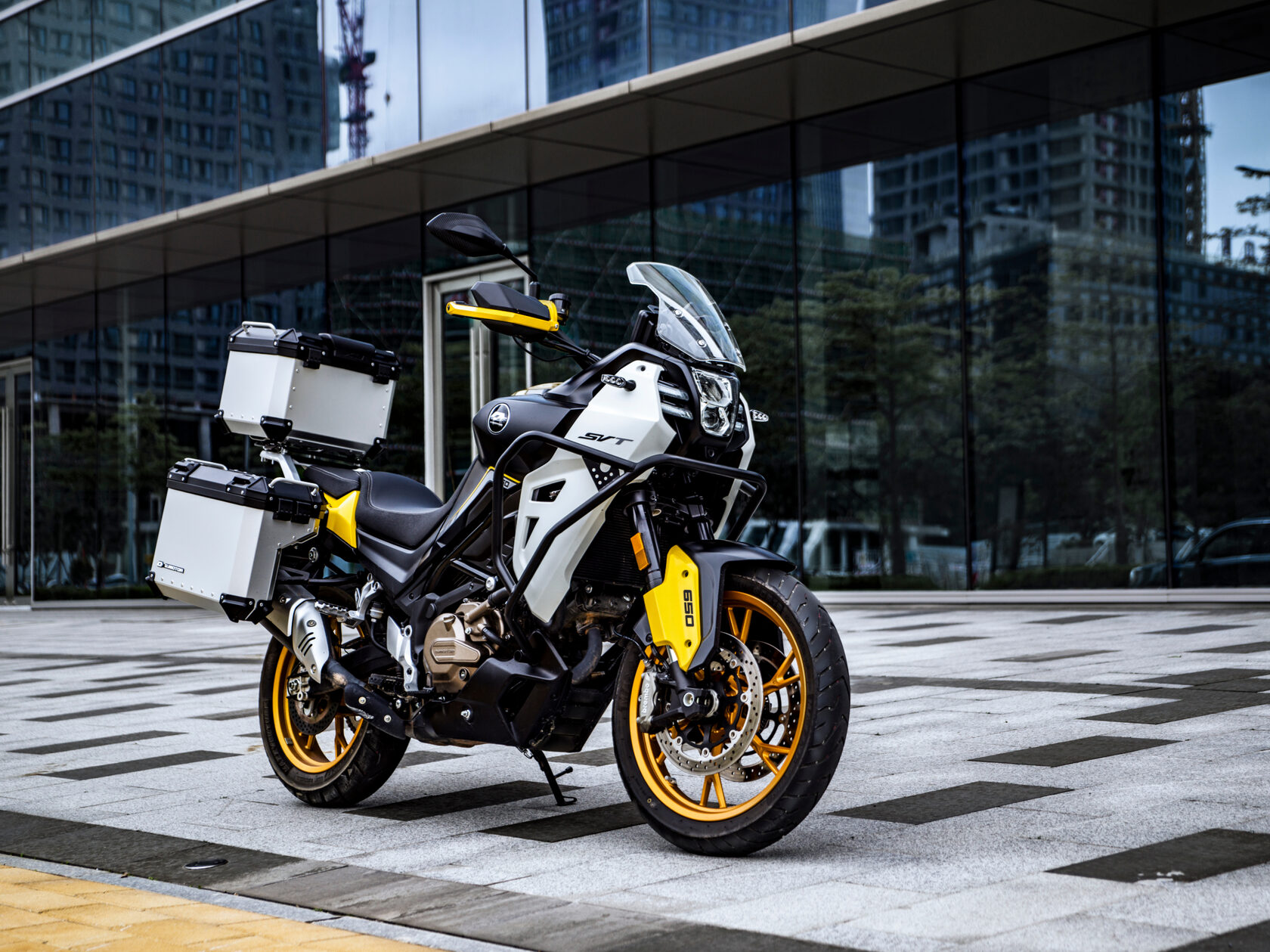 Weiß-schwarzer QJMOTOR SVT 650 X mit goldfarbenen Rädern und drei Aluminiumkoffern, vollständig ohne Fahrer auf einem Platz geparkt. Das Motorrad steht schräg zur Kamera auf gemusterten Betonplatten vor einem modernen Gebäude mit Glasfassade.