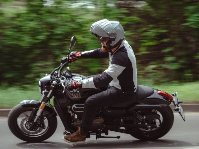 Schwarzer QJMOTOR SRV 12 in Fahrt, vollständig von der Seite vor verschwommenen grünen Bäumen aufgenommen. Die Person trägt einen silberfarbenen Helm, eine Motorradjacke und braune Stiefel und sitzt mit den Händen am Lenker auf dem Cruiser.