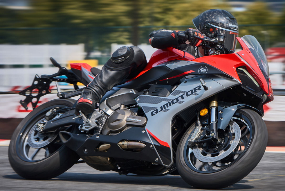 Rot-silber-schwarzer QJMOTOR SRK 600 RS während der Fahrt auf dem Asphalt einer Rennstrecke, schräg von vorn aufgenommen. Die Person trägt schwarzen Helm und dunkle Schutzkleidung; Strecke und Randsteine erscheinen hinter dem vollständig sichtbaren Motorrad unscharf.