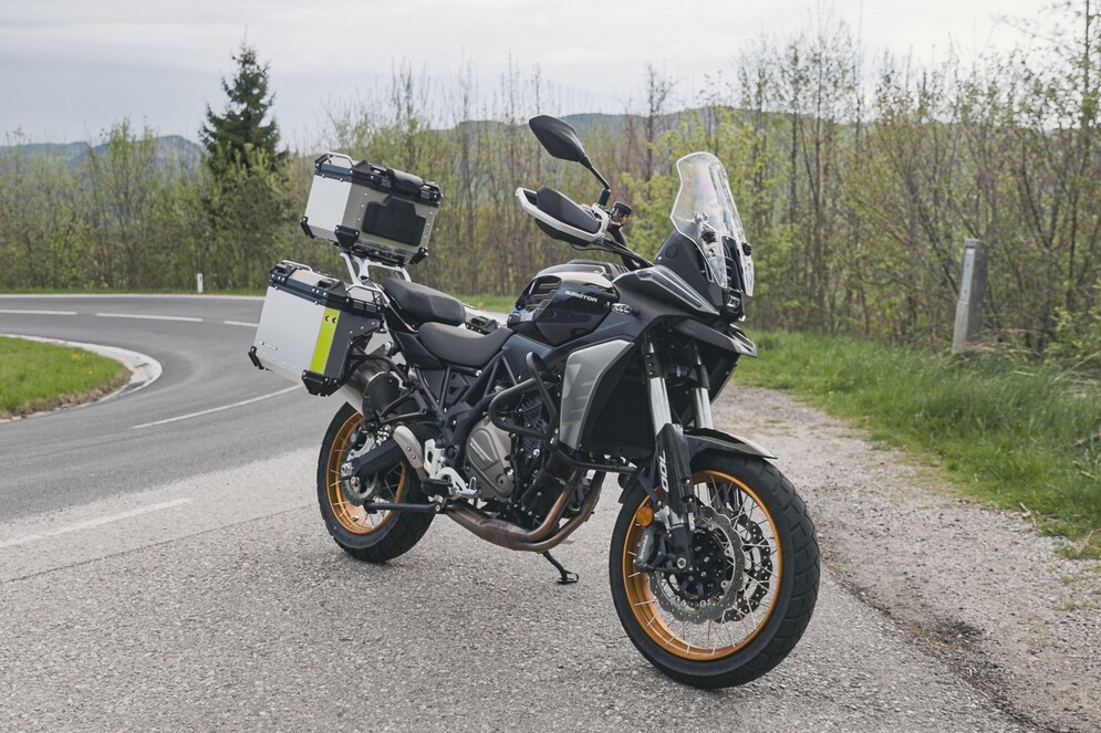 Grau-schwarzer QJMOTOR SRT 700 SX mit goldfarbenen Speichenrädern und Koffern, vollständig ohne Fahrer am Rand einer kurvigen Waldstraße geparkt. Asphalt und Schotter treffen unter dem Motorrad aufeinander; grüne Bäume und Gras liegen im Hintergrund.