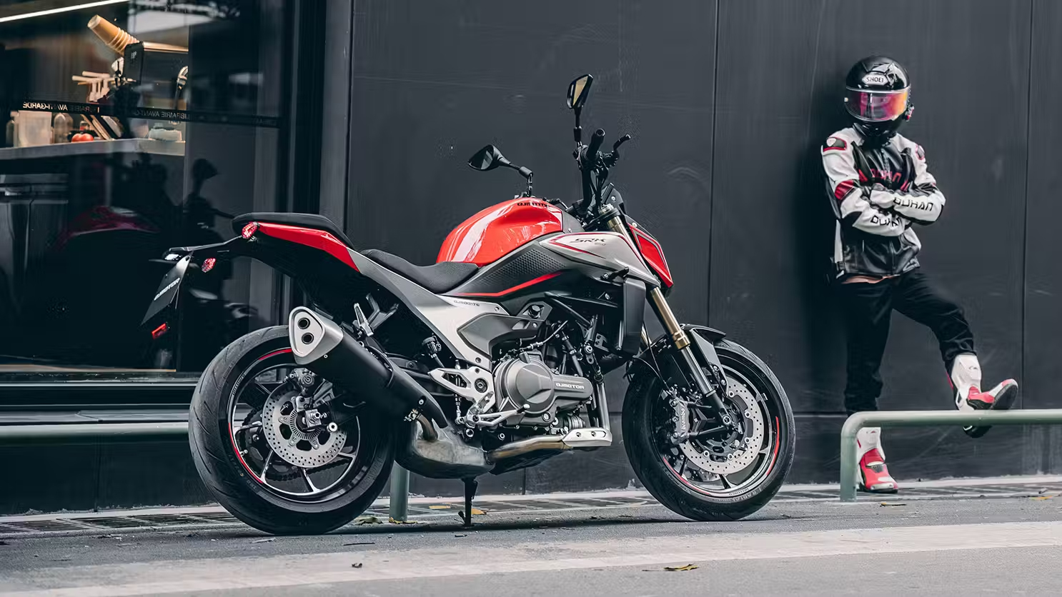 Rot-schwarzer QJMOTOR SRK 600 PRO, vollständig von der Seite auf einem modern gepflasterten Platz geparkt. Eine Person mit Helm und rot-schwarz-weißer Motorradkleidung steht im Vordergrund beim Vorderrad und lehnt an einer Wand.