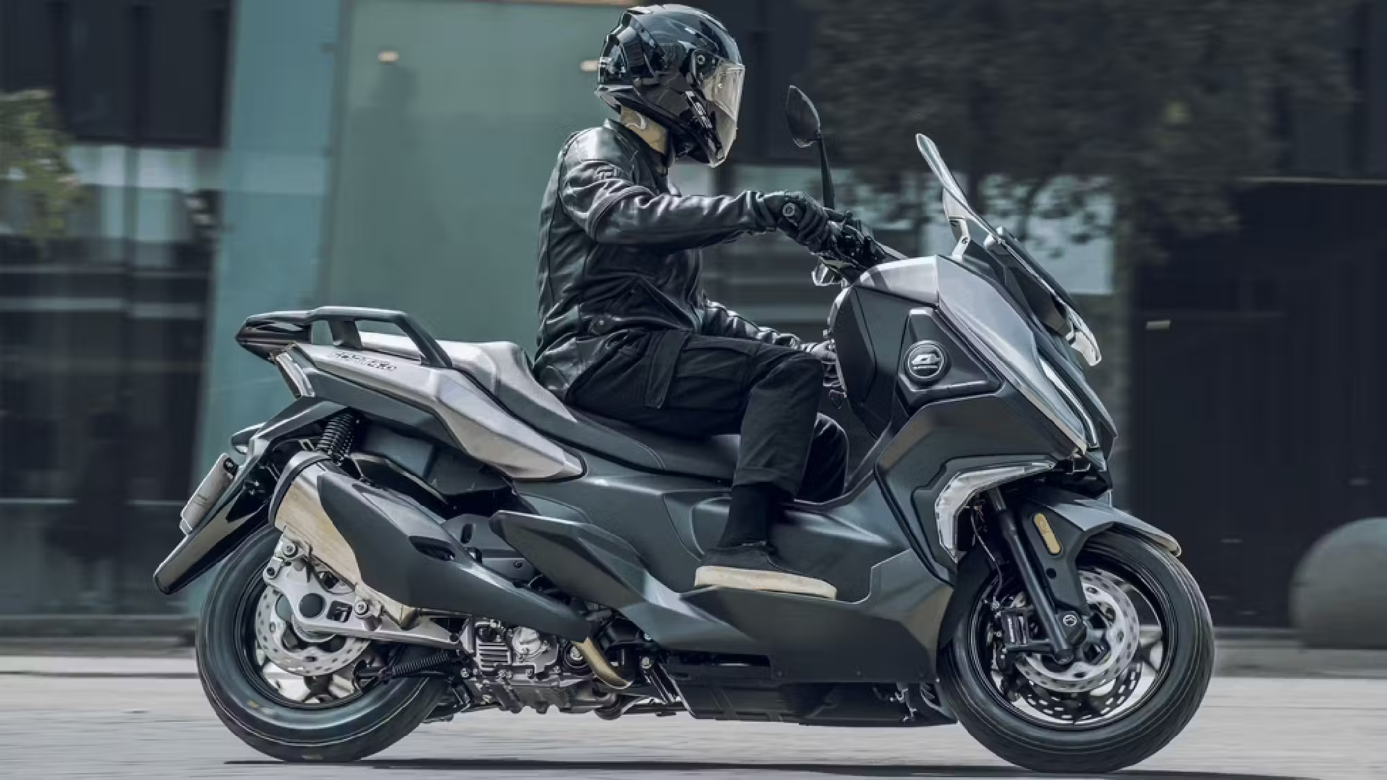 Grauer QJMOTOR FORT 4.0 in Fahrt auf einer städtischen Asphaltstraße vor einem Gebäude mit Glasfassade. Die Person trägt schwarzen Helm und dunkle Motorradkleidung; der Roller ist von der Seite vollständig sichtbar, der Hintergrund verwischt durch die Bewegung.