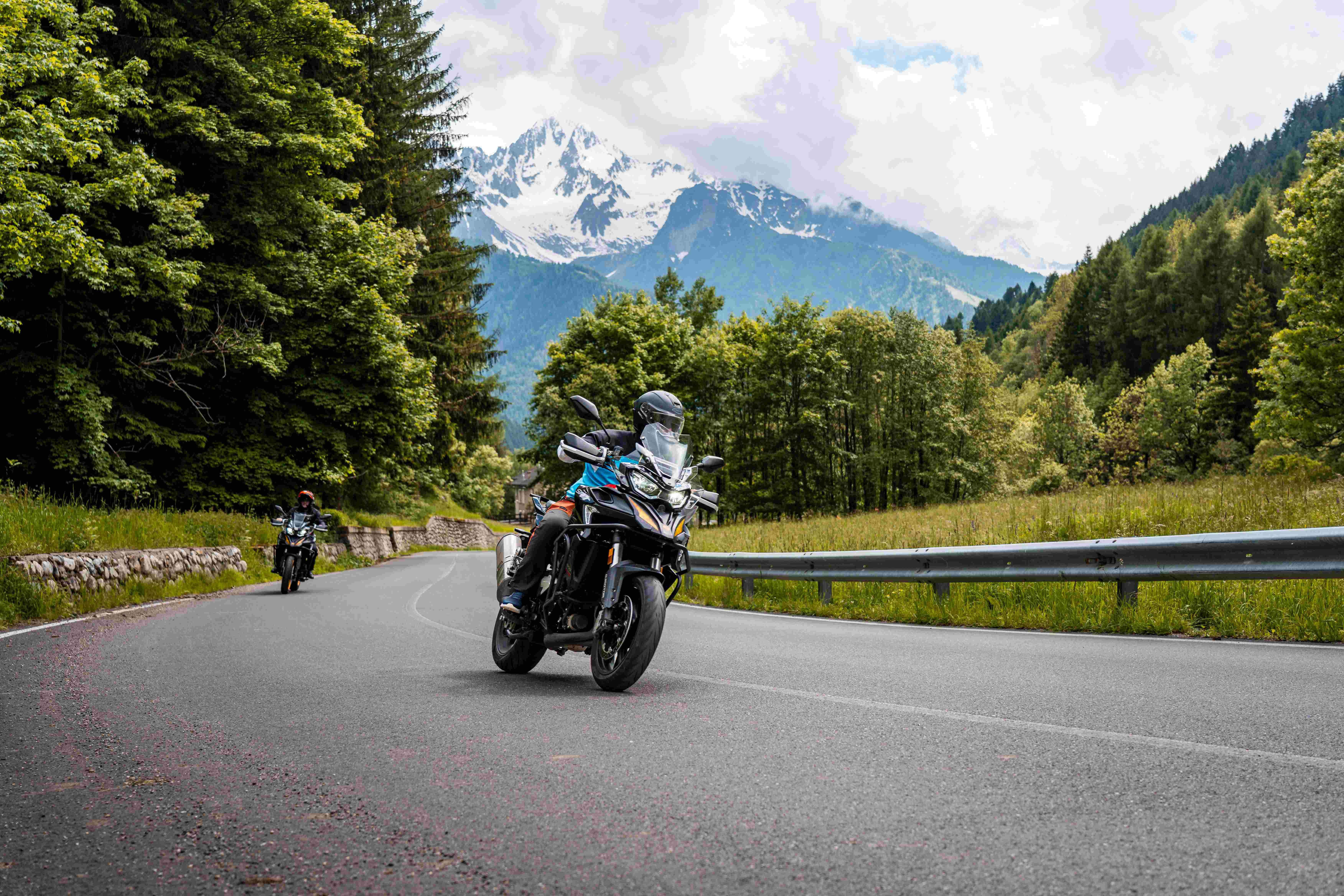Grau-schwarzer QJMOTOR SRT 900 SX mit Koffern während der Fahrt auf einer kurvigen Bergstraße. Die Person trägt Helm und grau-schwarze Schutzkleidung; Leitplanke, Nadelbäume und ein schneebedeckter Gipfel liegen dahinter, ein weiteres Motorrad folgt in großer Entfernung.