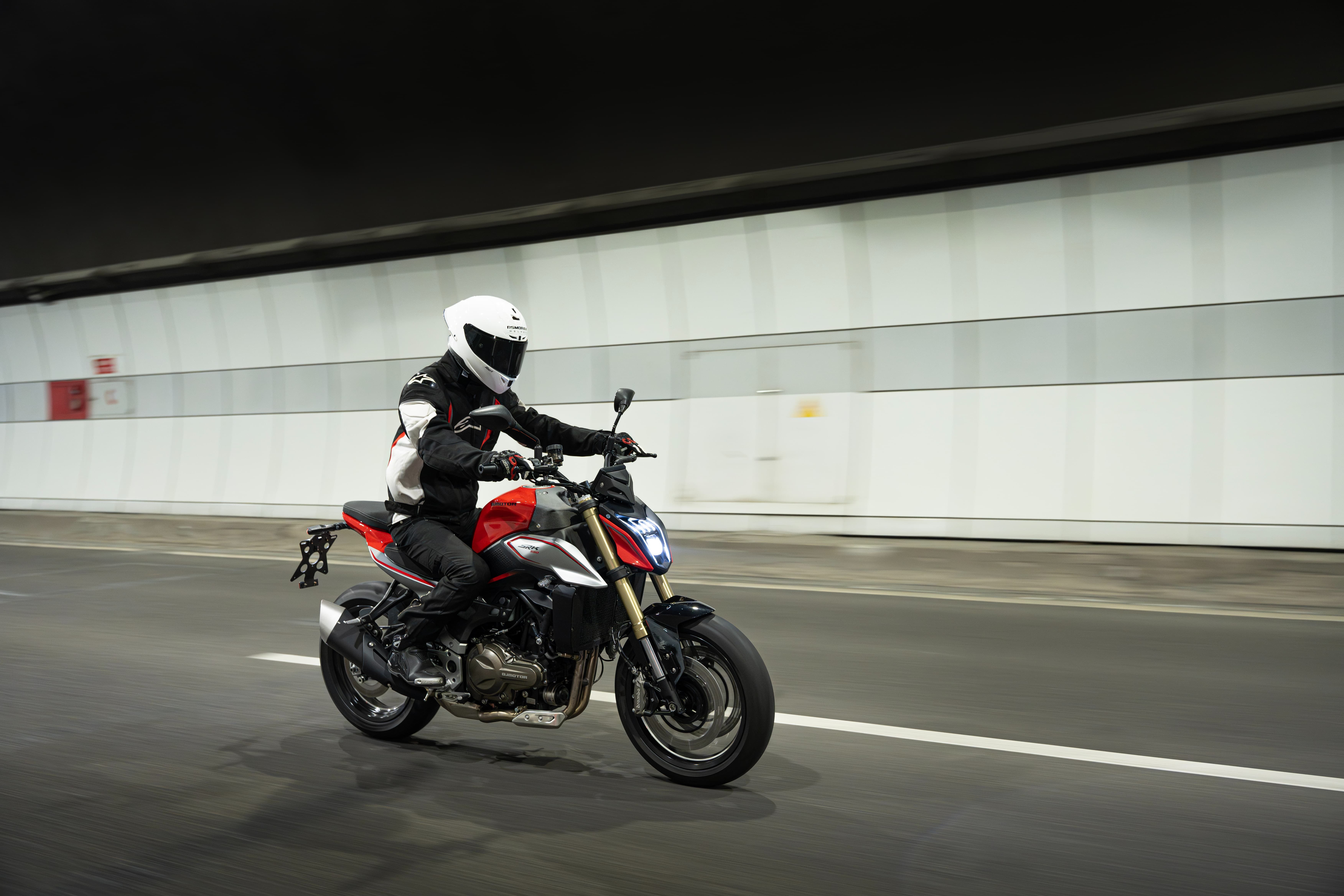 Rot-schwarzer QJMOTOR SRK 600 PRO in Fahrt durch einen beleuchteten Stadttunnel. Die Person trägt weißen Integralhelm und eine rot-schwarz-weiße Motorradjacke; das Motorrad ist von der Seite vollständig sichtbar, Tunnelwände und Lichter verwischen im Hintergrund.