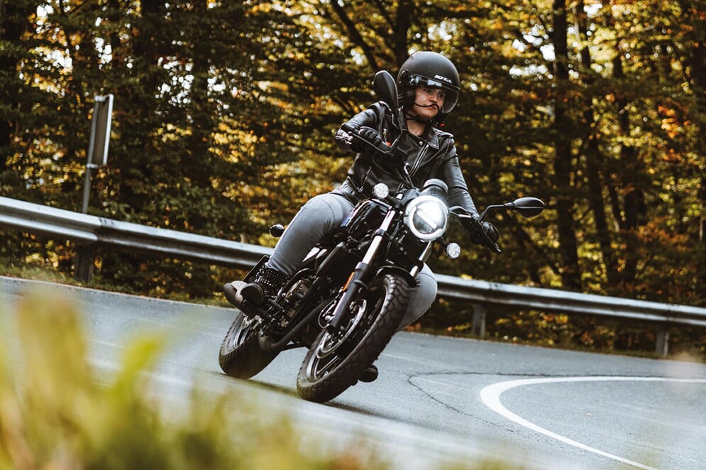 Schwarzer QJMOTOR SRV 125 mit einer Person in schwarzem Helm, dunkler Jacke und heller Hose während einer Kurvenfahrt. Die Aufnahme zeigt den Cruiser schräg von vorn; eine Leitplanke und herbstlich gefärbter Wald säumen die Straße.