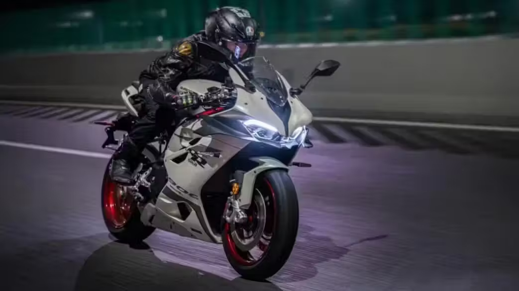 Weiß-schwarzer QJMOTOR SRK 921 RR mit roten Felgen während der Fahrt auf einer beleuchteten Asphaltstrecke bei Dunkelheit. Die Person trägt einen geschlossenen Helm und dunklen Rennanzug; Streckenmarkierungen und eine Begrenzungsmauer verlaufen hinter dem Motorrad.