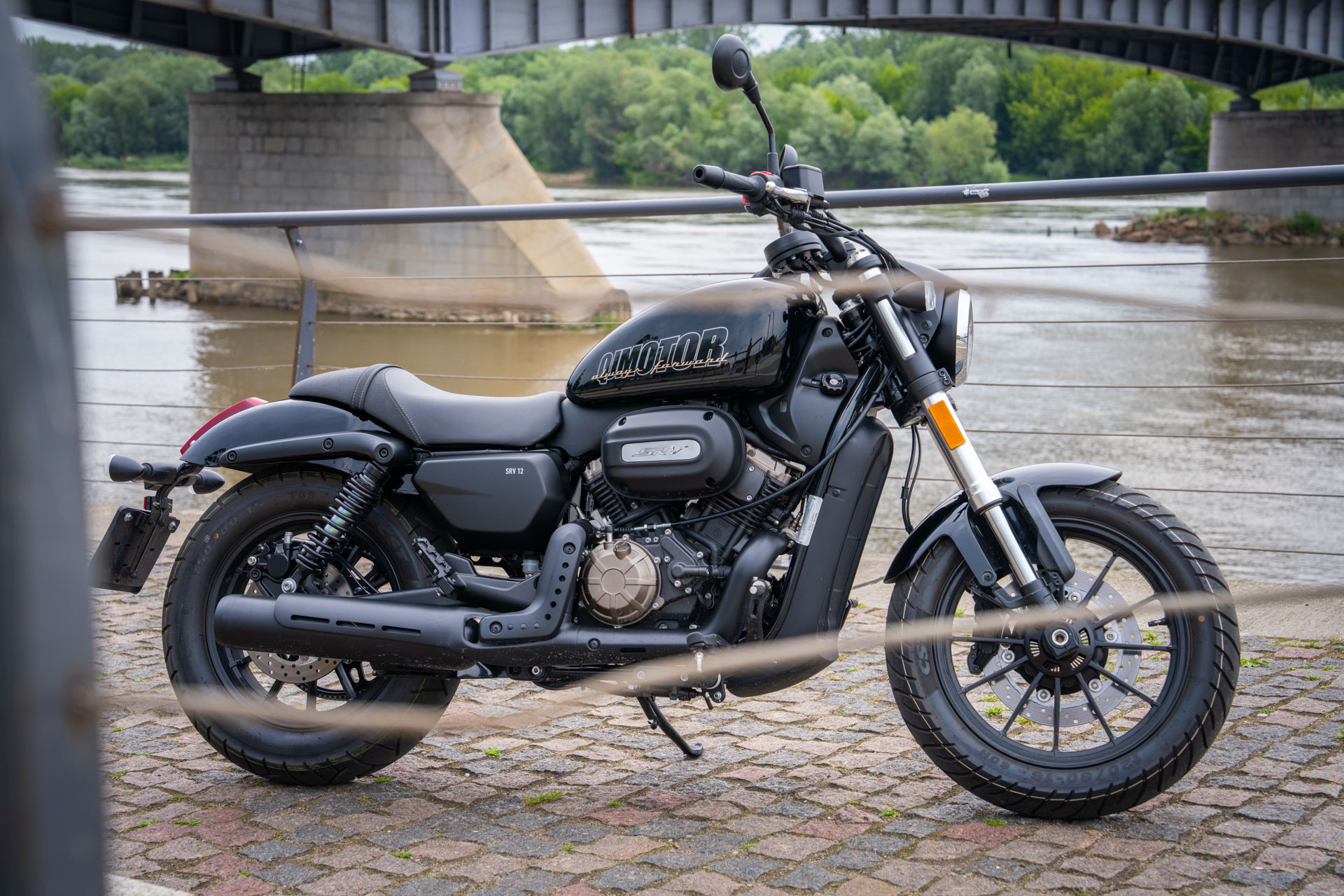 Schwarzer QJMOTOR SRV 12 ohne Fahrer, vollständig auf einer gepflasterten Uferpromenade geparkt. Hinter dem Cruiser verlaufen ein Metallgeländer und ein Fluss; eine große Stahlbrücke ist im Hintergrund zu sehen.