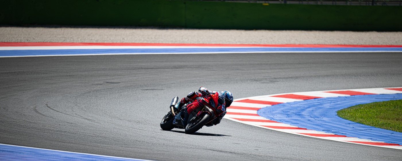 Rot-schwarzer QJMOTOR SRK 125 R mit einer Person in Rennanzug und geschlossenem Helm in deutlicher Schräglage auf einer Rennstrecke. Die weite Aufnahme zeigt Asphalt, Streckenbegrenzungen und blau-rot-weiße Randsteine rund um das Motorrad.