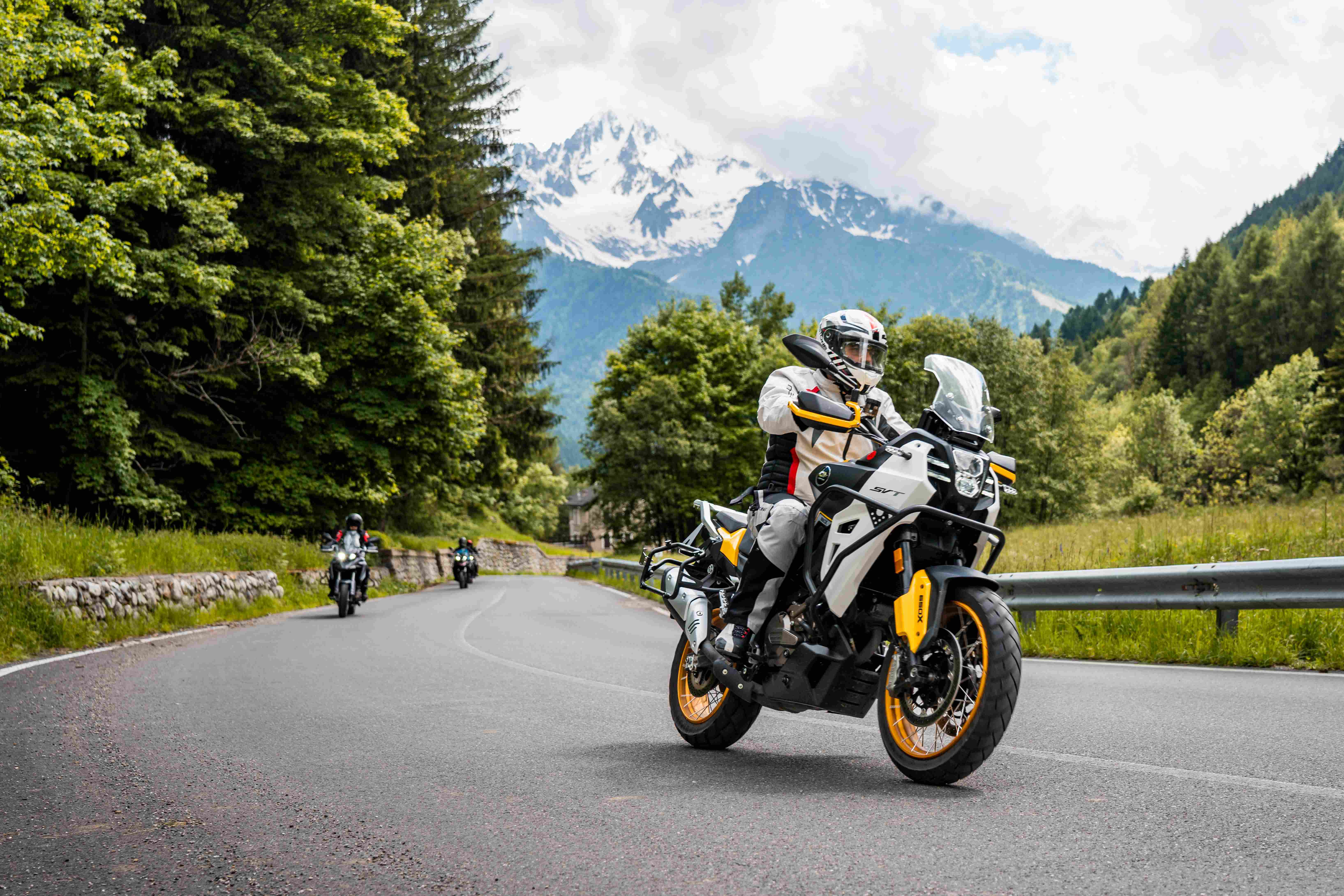 Weiß-schwarzer QJMOTOR SVT 650 X mit goldfarbenen Speichenrädern und Aluminiumkoffern in Fahrt auf einer Bergstraße. Die Person trägt weißen Helm und helle Schutzkleidung; Leitplanke, Nadelwald und schneebedeckte Berge liegen dahinter, ein weiteres Motorrad ist weit entfernt.
