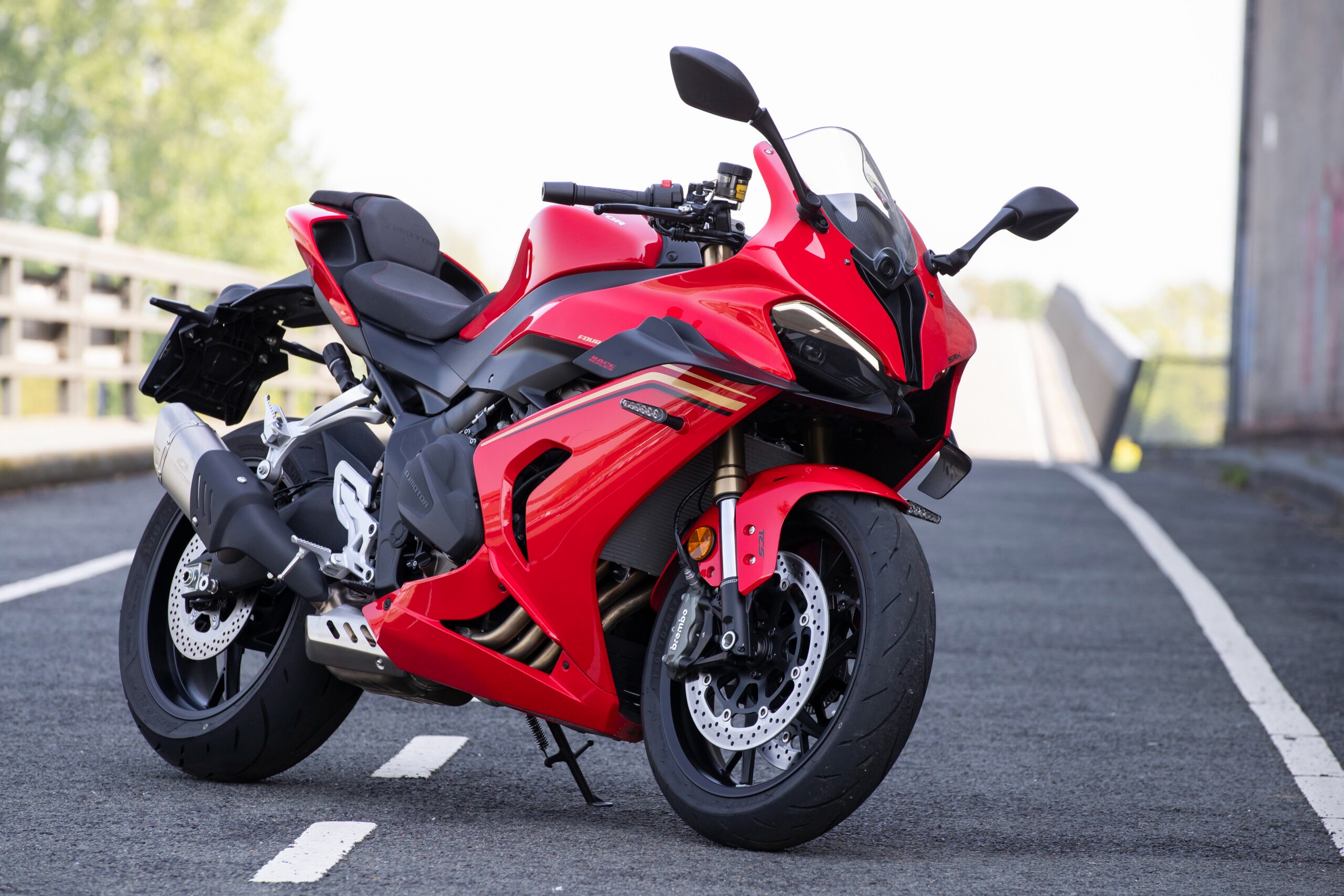 Roter QJMOTOR SRK 800 RR, vollständig ohne Fahrer schräg von vorn auf Asphalt geparkt. Die Aufnahme zeigt Verkleidung, Scheibe und beide Räder; ein helles Gebäude und Bäume stehen im Hintergrund.