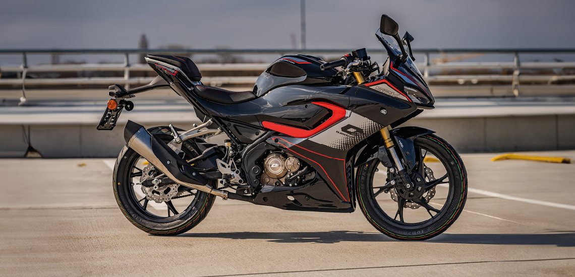 Schwarz-grau-roter QJMOTOR SRK 125 R, vollständig und ohne Fahrer auf dem Asphalt eines Fahrerlagerbereichs geparkt. Die seitliche Dreiviertelansicht zeigt Verkleidung, Sitzbank und beide Räder; Absperrungen und Streckenanlagen liegen dahinter.