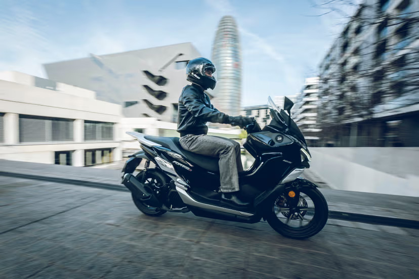 Grauer QJMOTOR FORT 125 N während der Fahrt auf einer Stadtstraße vor moderner heller Architektur. Die Person trägt einen Helm, eine dunkle Jacke und eine hellbraune Hose; beide Räder des Rollers sind sichtbar, der Hintergrund ist durch die Bewegung unscharf.