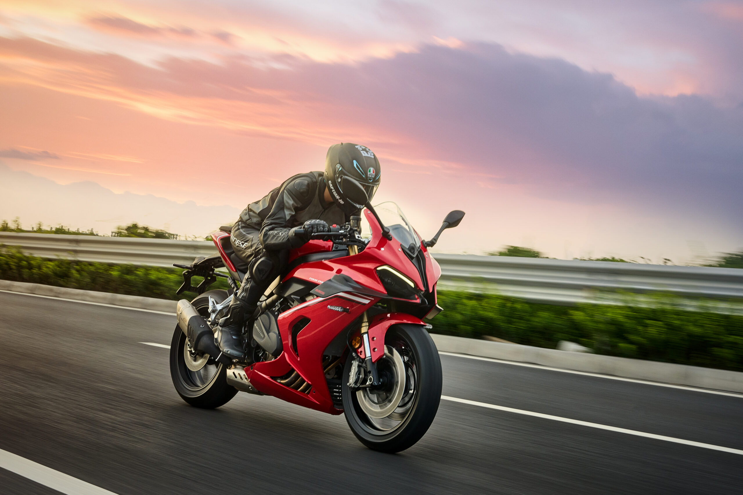 Roter QJMOTOR SRK 800 RR während der Fahrt auf einer breiten Asphaltstrecke vor einem rosafarbenen Abendhimmel. Die Person trägt schwarzen Helm und dunkle Motorradkleidung; Leitplanke und Hintergrund verwischen, das gesamte Sportmotorrad ist sichtbar.