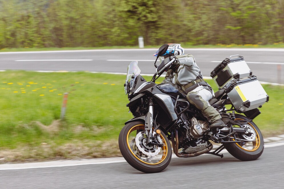 Grau-schwarzer QJMOTOR SRT 700 SX mit goldfarbenen Speichenrädern und Koffern während einer Kurvenfahrt auf einer Landstraße. Die Person trägt Helm und graue Schutzkleidung; grüner Straßenrand und Wald umgeben das vollständig sichtbare Motorrad.