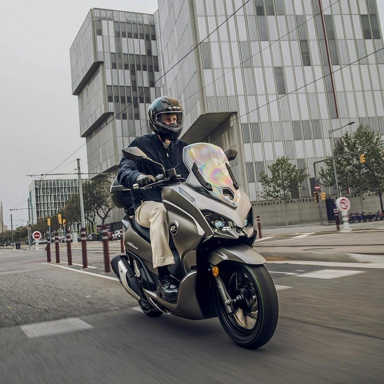 Silberfarbener QJMOTOR MTX 125 während der Fahrt durch eine moderne städtische Kreuzung. Die Person am Lenker trägt Helm, dunkle Jacke und helle Hose; Fahrbahnmarkierungen und Gebäude mit Glasfassaden umgeben den vollständig sichtbaren Roller.