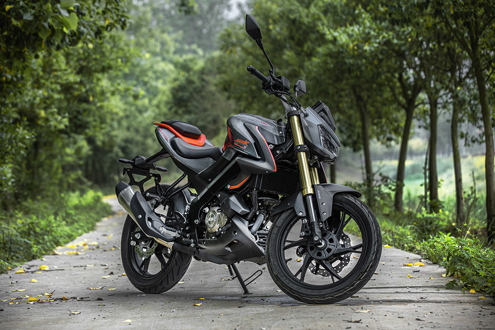 Dunkelgrau-schwarzer QJMOTOR SRK 125 S mit orangefarbenen Akzenten, vollständig schräg von vorn am bewaldeten Straßenrand geparkt. Der Asphalt liegt im Schatten grüner Bäume; das Motorrad steht allein, ohne Fahrer oder weitere Personen.