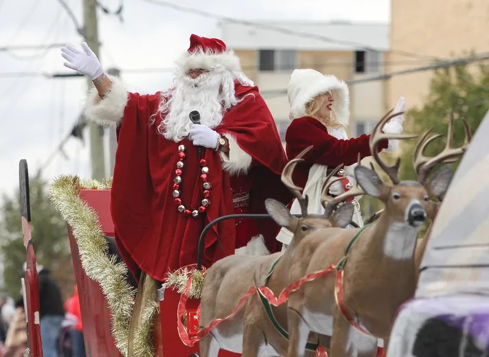 Santa and a woman in red and white costumes wave from a sleigh pulled by reindeer in a festive parade. Background shows city buildings.