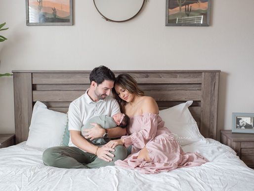 Couple lovingly cradles newborn on bed in cozy, neutral-toned room with framed cacti art and plant. Soft, serene mood at an in-home newborn session with a top newborn photographer in Surprise, Arizona.