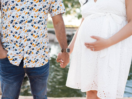 Pregnant woman in white dress and man in patterned shirt hold hands by a pond. Her hand rests on her belly, displaying a ring. Photographed by a Scottsdale Newborn Photographer.