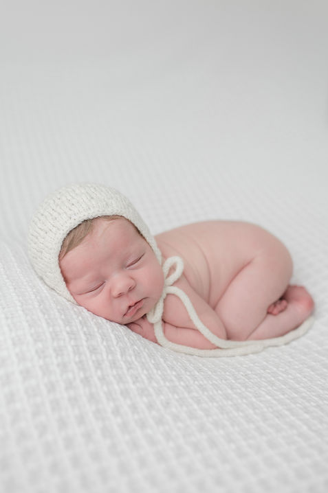 Newborn baby girl lays on white backdrop with white bonnet, at her in-home newborn session in Litchfield Park