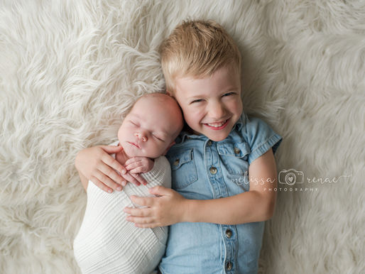 Smiling child in denim hugs a swaddled sleeping baby on a fluffy white surface. Calm and joyful mood at an in-home newborn session in Surprise, AZ. Text: "Melissa Renae Photography".