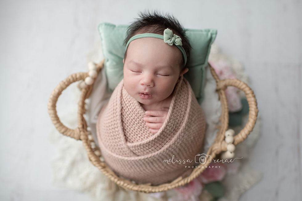 Sleeping baby in a basket, wrapped in a pink knit blanket. Green pillow and bow; calm setting at an in-home newborn session with a top newborn photographer in Surprise, Arizona.