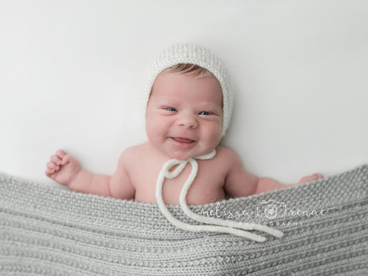 Smiling baby wearing a white knit bonnet, wrapped in a gray blanket, on a soft white background. Text: melissa renae photography. Post is to help planning your Phoenix newborn session.