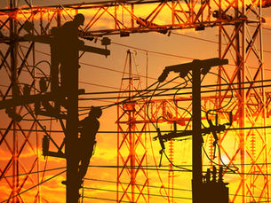 Silhuetas de trabalhadores de serviços públicos em postes de energia e torres de transmissão elétrica contra um céu alaranjado e amarelo ao pôr do sol.