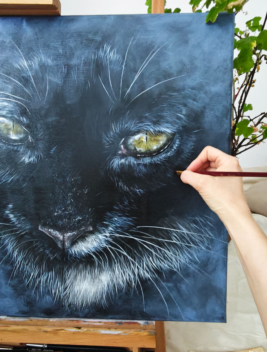 Nahaufnahme einer Katzenporträt-Malerei auf Leinwand; eine Hand setzt mit Pinsel Details am Auge, dunkler, konzentrierter Arbeitsmoment.