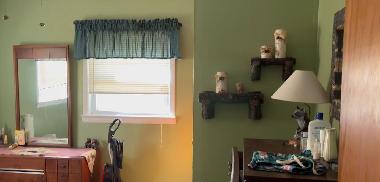 A bedroom with soft green walls, a wooden dresser with a mirror, and rustic log-style wall shelves holding decorative candles. A vacuum and personal items sit nearby.