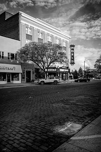 Black and white photograph of The Bricks in downtown Kearney, Nebraska, featuring the World Theatre marquee, historic storefronts, and a cobblestone street with a small reflective puddle in the foreground.