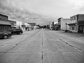 Black-and-white photograph of downtown Neligh, Nebraska, showing an empty main street lined with small businesses, parked cars, and overcast skies with dramatic clouds.