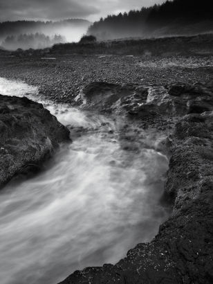 A misty coastal stream flows through dark volcanic rocks toward a pebble-strewn shore, with fog drifting along forested bluffs beneath a moody, overcast sky.