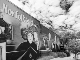 Black-and-white photograph of the Johnny Carson mural in downtown Norfolk, Nebraska, featuring painted portraits and the words "Norfolk's Own" on the side of a downtown building beneath a cloud-filled sky.