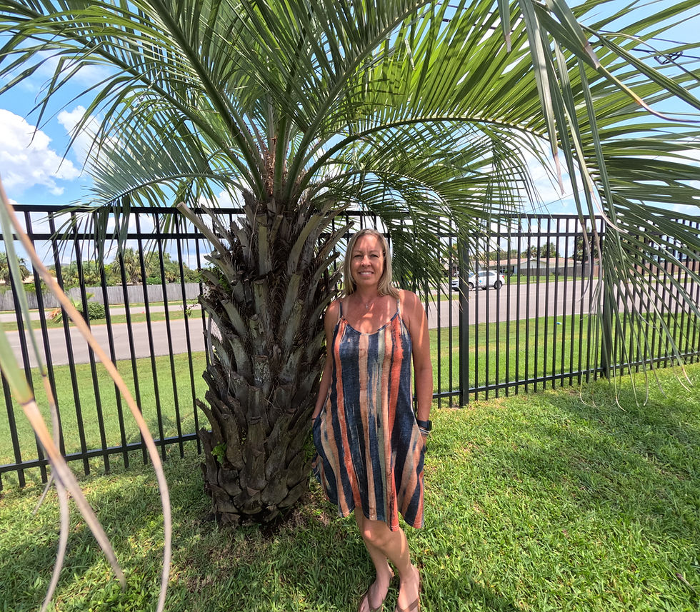 A real estate agent standing in front of a palm tree and black iron fence on a sunny day in Fort Walton Beach, Florida, representing a friendly and approachable local expert specializing in military relocation and coastal properties.